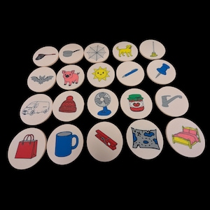Può includere: Una collezione di 24 gettoni rotondi in legno, ognuno con un'illustrazione disegnata a mano. Le immagini includono un cane, un maiale, un sole, una borsa della spesa rossa, una tazza blu e un letto. I gettoni sono disposti su una superficie nera.