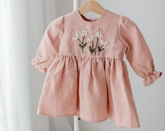 May include: A pale pink linen dress with embroidered white daisies on the bodice. The dress has long sleeves with ruffles at the cuffs and a gathered skirt.