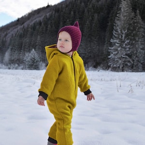 May include: A young child wearing a yellow snowsuit and a burgundy knitted hat walks through a snowy field. The child is looking to the right of the frame.
