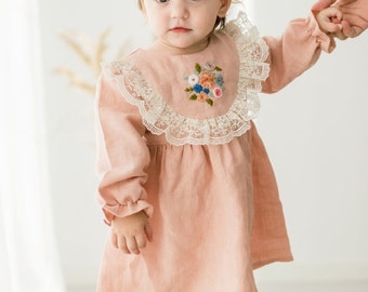May include: A little girl wearing a pink linen dress with a white lace collar and embroidered flowers. The dress has long sleeves with ruffles at the cuffs.