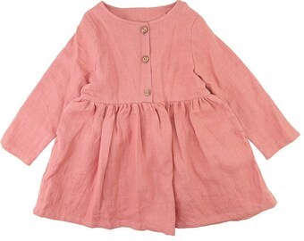 May include: A light pink long-sleeved dress with a round neckline and a gathered skirt. The dress features three wooden buttons down the front and is made of a soft, textured fabric. The dress is designed for children.