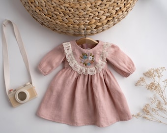 May include: A pink linen dress with a lace trim and embroidered flowers. The dress has long sleeves and a gathered skirt.