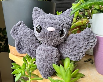 Puede incluir: Un juguete de peluche de murciélago de crochet gris con ojos negros y una nariz rosa. El juguete está sentado sobre una superficie de madera con plantas verdes.