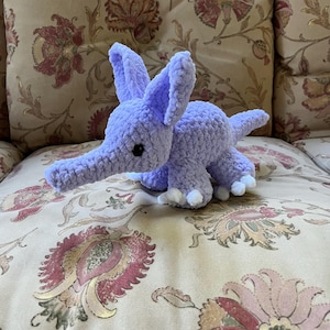 Può includere: Un elefante all'uncinetto lavanda fatto a mano, con grandi orecchie, una lunga proboscide e piccoli piedi bianchi. Il peluche è su un cuscino a fantasia floreale. L'elefante è un giocattolo morbido e coccoloso.