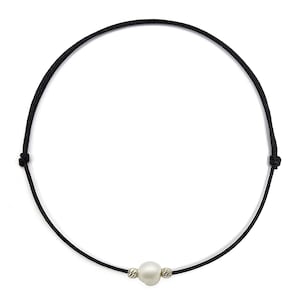 Op de afbeelding: Een zwarte koordketting met een enkele, grote, witte parel. De parel wordt geflankeerd door twee kleine, getextureerde zilveren kralen. De ketting heeft een eenvoudig en elegant ontwerp.