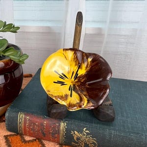 May include: A brown ceramic pot with a trailing green plant sits next to a decorative flower-shaped dish. The dish is yellow and brown, resting on a dark wooden stand. A vintage book is in the background.