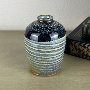 May include: A small ceramic bud vase with a narrow neck and a rounded body. The upper portion features a glossy dark blue and black speckled glaze, while the lower section displays a textured, ribbed surface with a muted grey and brown finish.