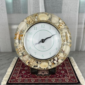 May include: A round analog clock with a clear resin frame embedded with various tan, white, and brown seashells and a small starfish. The clock face is white with black numerals and hands, featuring the text Sands of Time in the center.