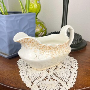 May include: A cream-colored ceramic gravy boat featuring a delicate, light brown floral pattern along the upper rim, a curved handle, and a wide pouring spout, resting on a white, intricate, heart-shaped crocheted lace doily.