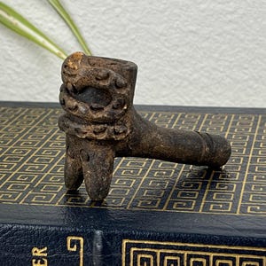 May include: A small, intricately carved wooden pipe with a lion-like head and a cylindrical stem. The pipe is dark brown and rests on a vintage book titled "The Odyssey of Homer". The book has a navy blue cover with gold lettering and a decorative pattern.