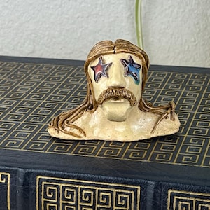 May include: A small, ceramic bust with a mustache and long hair, featuring star-shaped accents in the eyes. The bust is placed on a dark blue book titled "The Odyssey of Homer". The book has gold-colored geometric patterns.
