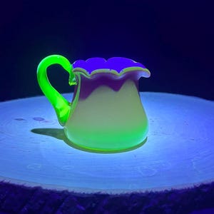 May include: A small, glowing glass pitcher with a handle, sitting on a wooden disc. The pitcher has a ruffled rim with purple and yellow accents. The handle and base of the pitcher glow bright green.