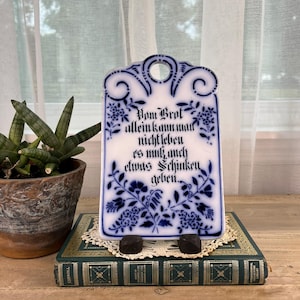 May include: A rectangular white ceramic wall plaque with rounded top edges and a hanging hole, featuring blue floral patterns and German text that reads: Vom Brot allein kann man nicht leben es muss auch etwas Schinken geben.