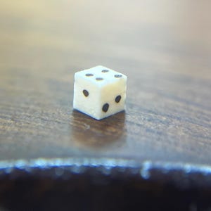 May include: A small, off-white die with black dots, resting on a wooden surface. The die is in sharp focus, with the background softly blurred. The image is a close-up shot, highlighting the details of the dice.