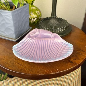 May include: A ceramic seashell-shaped dish in shades of lavender and light blue. The dish has a ribbed texture and a raised, rounded element at the top. It sits on a round, dark wooden table, with a lamp base and potted plants in the background.