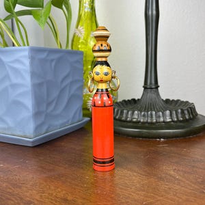 Peut inclure: Poupée en bois vintage avec un corps rouge et une tête empilée avec un visage peint et des créoles dorées. La poupée a un corps cylindrique avec des rayures noires et des pois rouges. La poupée mesure environ 15 cm de haut.