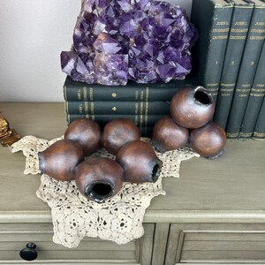 May include: A collection of brown, gourd-shaped pottery pieces with open tops, arranged on a cream-colored lace doily. A large purple amethyst crystal sits atop a stack of vintage books. A decorative figurine and a row of books are also visible.