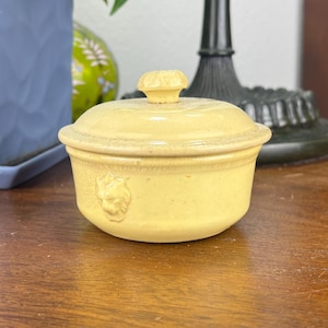 May include: A small, round, yellow ceramic dish with a lid. The dish has a decorative relief design on the side and a small knob on the lid. The dish is sitting on a wooden table.