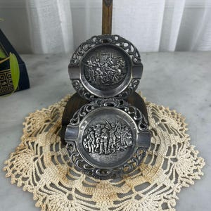 May include: Two circular silver-toned metal ashtrays featuring ornate, decorative borders and central embossed relief scenes. The top ashtray depicts a group of figures in an interior setting, while the bottom ashtray shows a group of figures outdoors.
