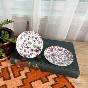 May include: A still life featuring a small potted plant with green leaves and red flowers, a round decorative dish with a floral pattern, and a small oval dish with a floral pattern. The dishes have gold trim.