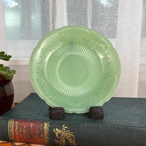 May include: A light green glass plate with a decorative rim sits on a dark green book. A small potted plant with red flowers and a brown and beige pot is to the left. The background includes a window with white curtains.
