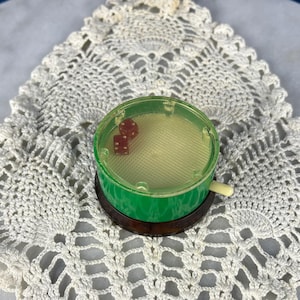 May include: A vintage dice shaker with two red dice inside, set on a brown base and a green top. The shaker sits on a white, crocheted doily with a detailed openwork pattern. The background is a marble surface against a blue backdrop.