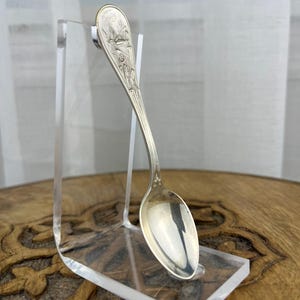 May include: A silver-toned metal spoon featuring an embossed design of a bird in flight and floral motifs on the handle. The spoon is displayed on a clear acrylic stand, showcasing its polished finish and detailed engraving on the upper handle.