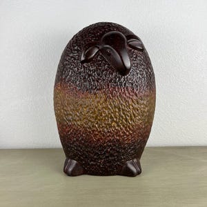 May include: A ceramic sheep figurine featuring a textured, wool-like surface with a gradient color scheme transitioning from dark brown at the top and bottom to a lighter amber-brown band across the center, with smooth, dark brown ears and feet.