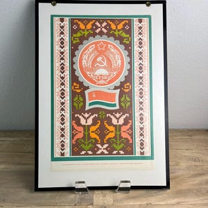May include: A framed poster featuring a brown background with a central circular emblem displaying a hammer and sickle, a star, and the text LTSR. Below the emblem is a flag with red, white, and green stripes, surrounded by geometric floral patterns.