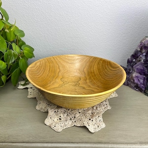 May include: A light brown wooden bowl with a natural wood grain pattern. The bowl is round with a slightly flared rim and sits on a cream-colored lace doily. The bowl is empty and appears to be new.
