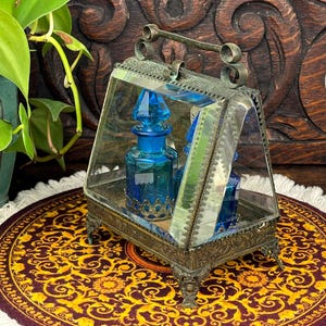 May include: An antique glass display case with a hinged top, housing two cobalt blue perfume bottles. The case has ornate metalwork and sits on a decorative, patterned mat. The background features a wooden carved design and a potted plant.