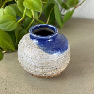 May include: A small, round ceramic vase with a textured, light grey body and a dark blue rim. The vase has a simple, minimalist design and is suitable for small flowers or decorative purposes. The vase is approximately 7.6 cm in diameter.