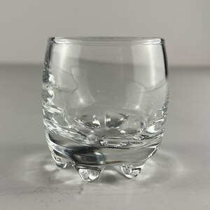 Peut inclure: Un petit verre clair et arrondi, avec une base épaisse et trois petits pieds. Le verre présente un motif texturé et tourbillonnant au milieu. Le verre est conçu pour contenir des liquides.