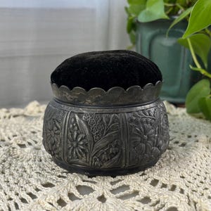 May include: An antique silver-toned pin cushion with a black velvet top. The cushion sits on a cream-colored, crocheted doily. The metal base is decorated with floral and leaf patterns. A green potted plant is in the background.