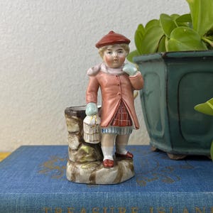 May include: A small, vintage figurine of a child in a red beret and pink coat, holding a basket. The figurine stands on a stone-like base. The background includes a blue book with gold lettering and a green plant in a dark green pot.