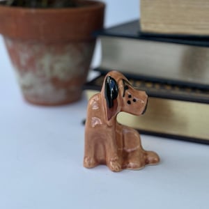 May include: A small, brown ceramic dog figurine with black ears and spots. The dog is in a seated position. A potted plant and a stack of books are in the background.