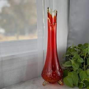 May include: A tall, slender, red glass vase with a flared, ruffled top and three small feet. The vase has a textured surface with small, round bumps. The top of the vase is a golden yellow color.