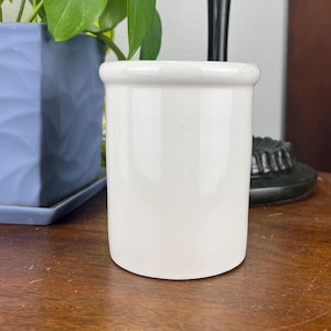 May include: A small, white ceramic jar with a rounded body and a slightly flared rim. The jar is smooth and glossy, and it sits on a wooden surface. The background includes a plant in a blue pot and a black lamp.