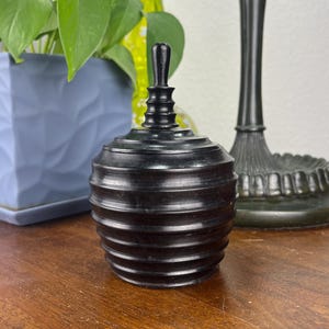May include: A black, wooden, lidded jar with a ribbed design. The jar sits on a wooden table, with a blue square planter with green plants and a green vase in the background. A black lamp is also visible.