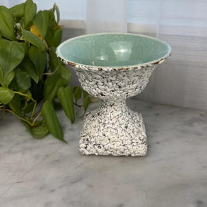 May include: A pedestal bowl with a speckled white and black exterior and a light green interior. The bowl has a wide, slightly flared rim and sits on a square base. The bowl is likely ceramic and is displayed on a marble surface.