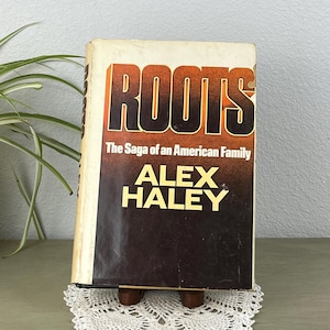 May include: A vintage copy of "Roots: The Saga of an American Family" by Alex Haley. The book has a worn cover with the title in orange and black. It rests on a small wooden stand atop a white lace doily, with a plant in the background.