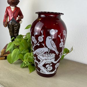 May include: A ruby red glass vase with a white etched design of birds on branches with flowers. The vase has a ruffled rim and a rounded body. A decorative lamp and a cat figurine are in the background.