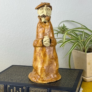 May include: A ceramic figurine of a man in a brown coat and beret. The figure has a mustache and is holding his hands together. The figurine is sitting on a book titled "The Odyssey of Homer".