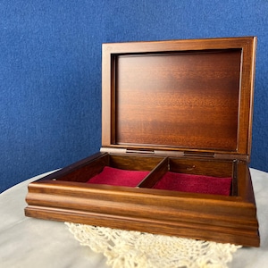 May include: Open wooden jewelry box with a rich, dark brown finish. The interior is lined with red velvet and divided into two compartments. The box is set against a blue background.