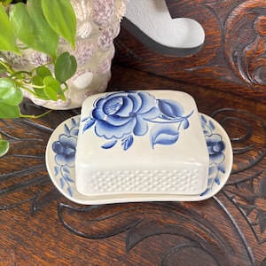 May include: A white ceramic butter dish with a blue floral design. The dish has a rectangular base and a matching lid, both decorated with blue roses and leaves. The dish sits on a matching oval plate.