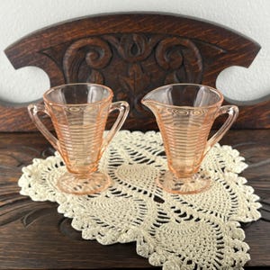 May include: Two vintage, light pink glass cream and sugar set. The sugar bowl has two handles, and the creamer has a single handle. Both pieces have a ribbed design. They sit on a cream-colored, crocheted doily, against a dark wood background.