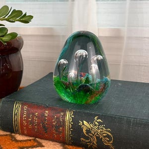 May include: A brown and cream ceramic pot with a flowering plant, a green and clear glass paperweight, and a vintage book with gold lettering sit on an orange patterned surface.