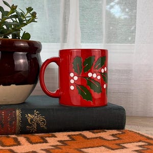 May include: A red ceramic mug with a green and white holly design sits on a dark green book next to a brown and cream-colored flower pot with a pink flower. The items are on a table with an orange patterned rug.