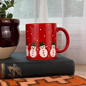 May include: A red mug with a snowman design sits on a vintage book next to a potted plant with a pink flower. The mug has a red handle and is decorated with white snowmen wearing black hats. The plant pot is brown with a cream-colored base.