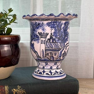 May include: A brown and cream-colored ceramic pot with a red flower and green plant sits next to a blue and white decorative vase. The vase features a detailed illustration of a building. Both items rest on a green book.
